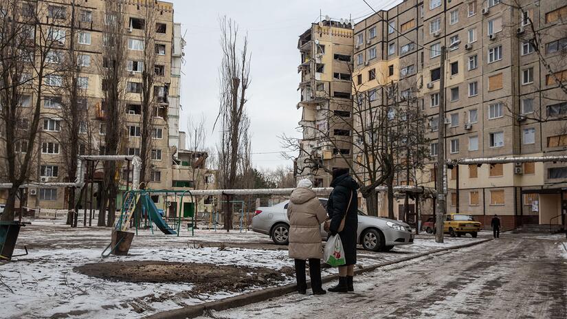 Two women stand in Dnipro, Ukraine, in the snow, looking at a gap where a building used to be before being destroyed during the conflict.
