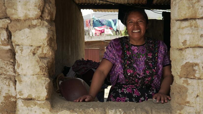 غلاديس غوميز تبتسم وهي تقف في منزلها مرتديةً اللون الأرجواني في زي ويبيل، وهو الزي التقليدي في غرب غواتيمالا. 