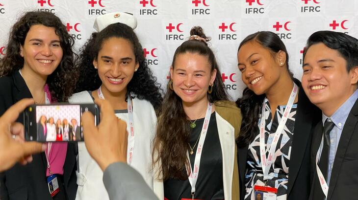 Youth volunteers at the General Assembly 2022 smile and pose for a picture in front of an IFRC branded photo wall.
