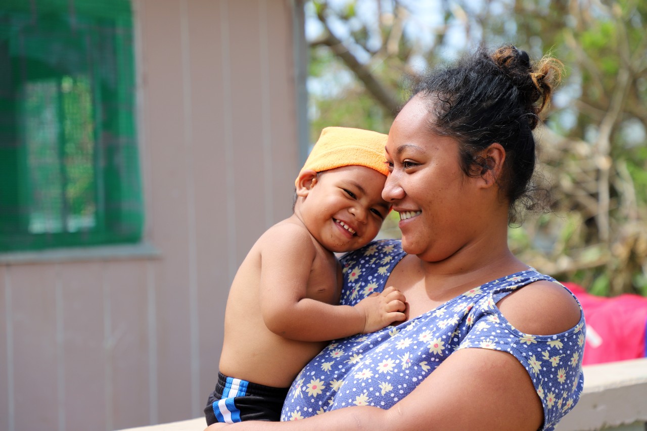 A mother in Tongatapu, Tonga stands with her baby boy nestled against her shoulder as she receives shelter assistance from Tonga Red Cross volunteers.