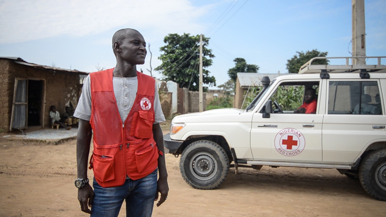 Nigerian Red Cross volunteer, Aliyu, wears his red vest and stands in front of a Nigerian Red Cross vehicle ready to help his community as they face food insecurity.