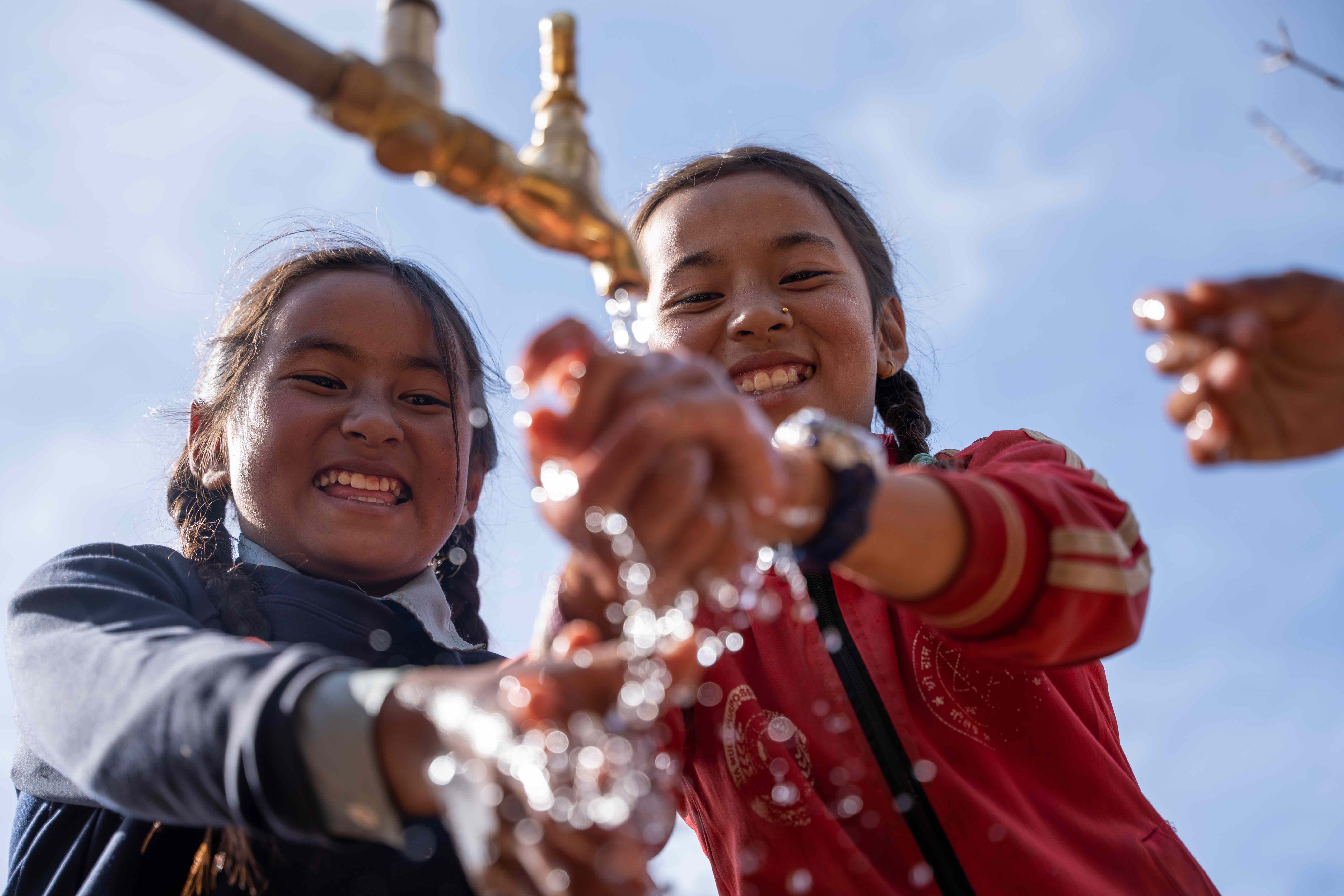 Two young children smile and laugh as they wash their hands in a newly built water tap in their village in rural Nepal.