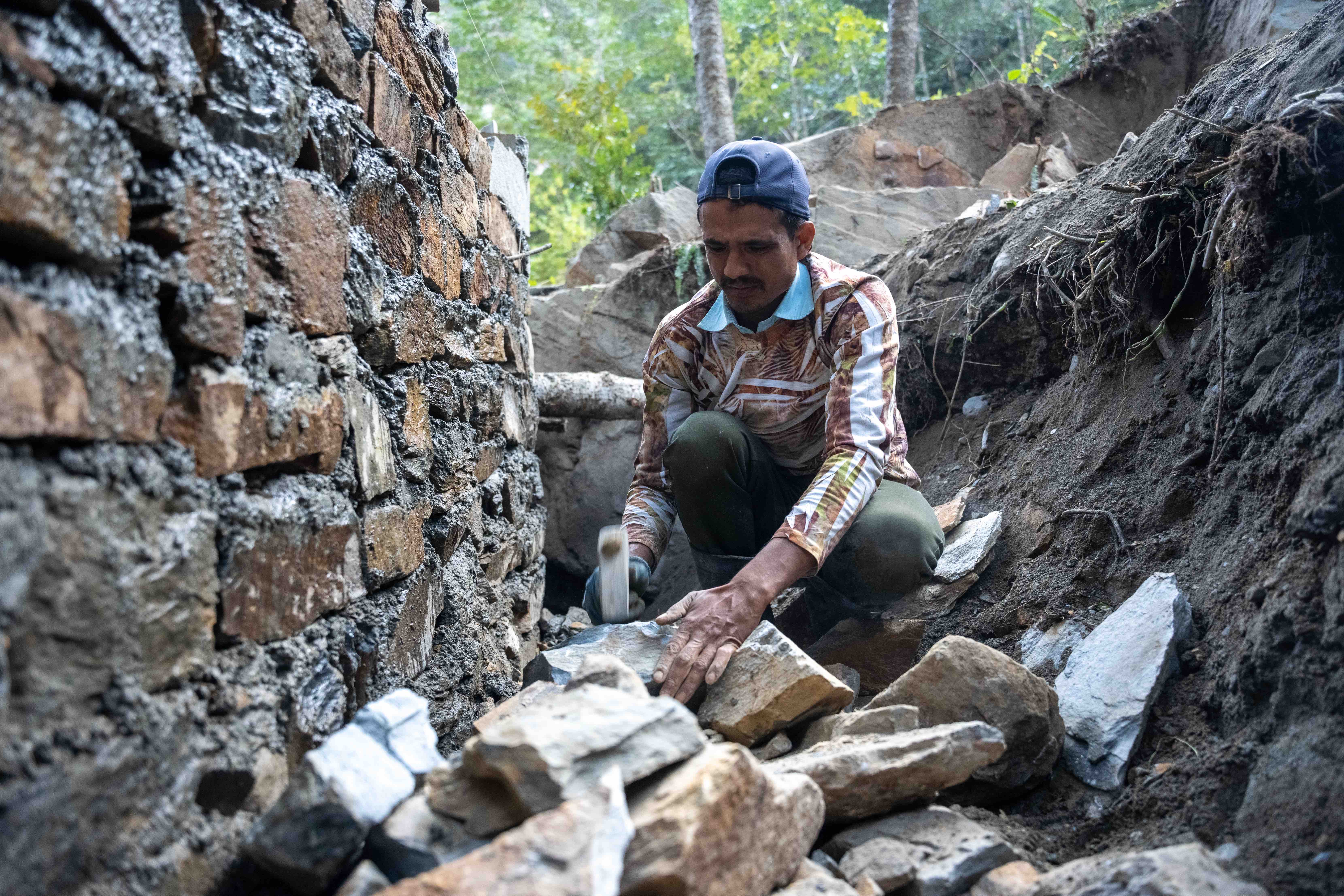 In one village, this man uses a hammer to shape rocks that will be used to build the wall of a water basin at a new water distribution point in rural eastern Nepal.