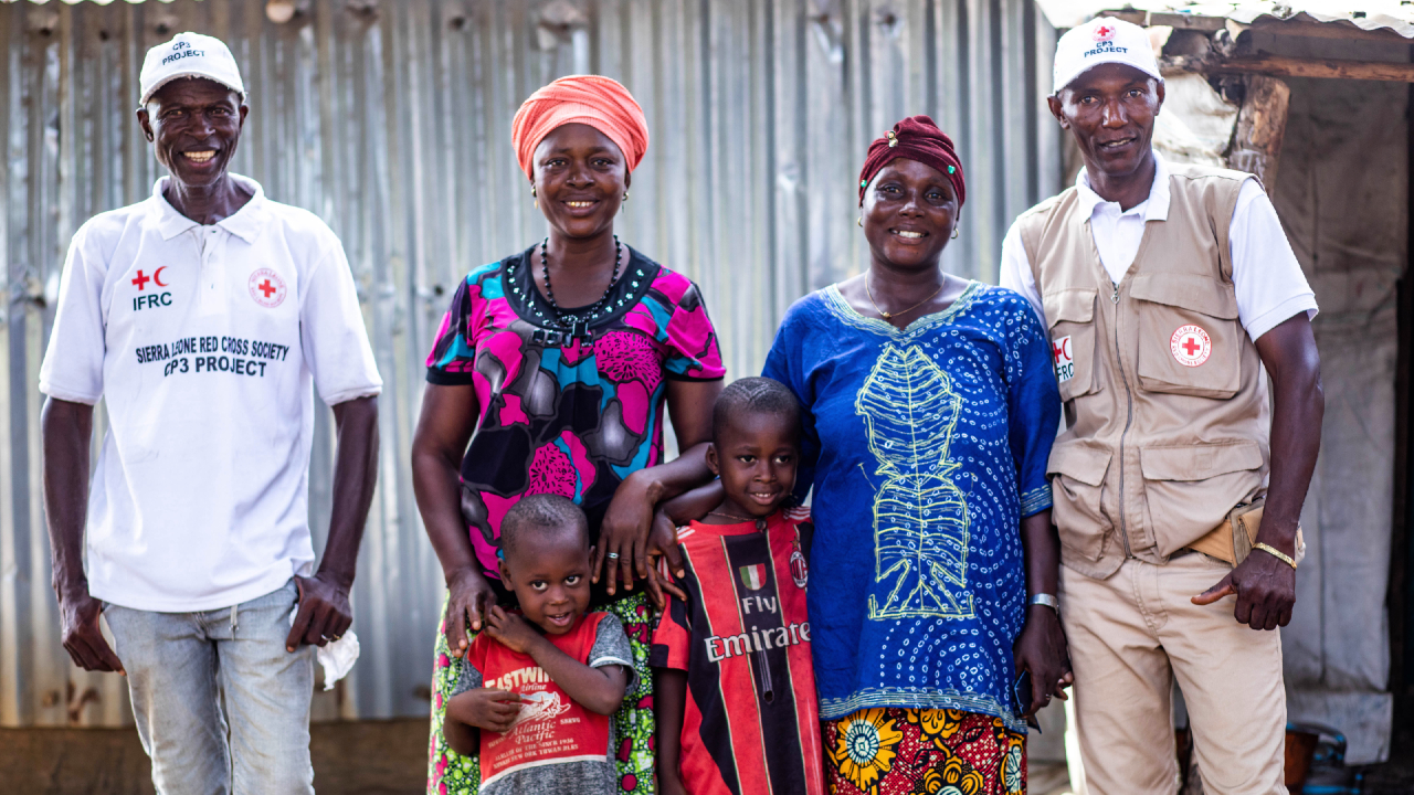 Sierra Leone Red Cross volunteer Momoh and supervisor Jobel stand with women and children from Makuma village who they supported during a measles outbreak in early 2022.