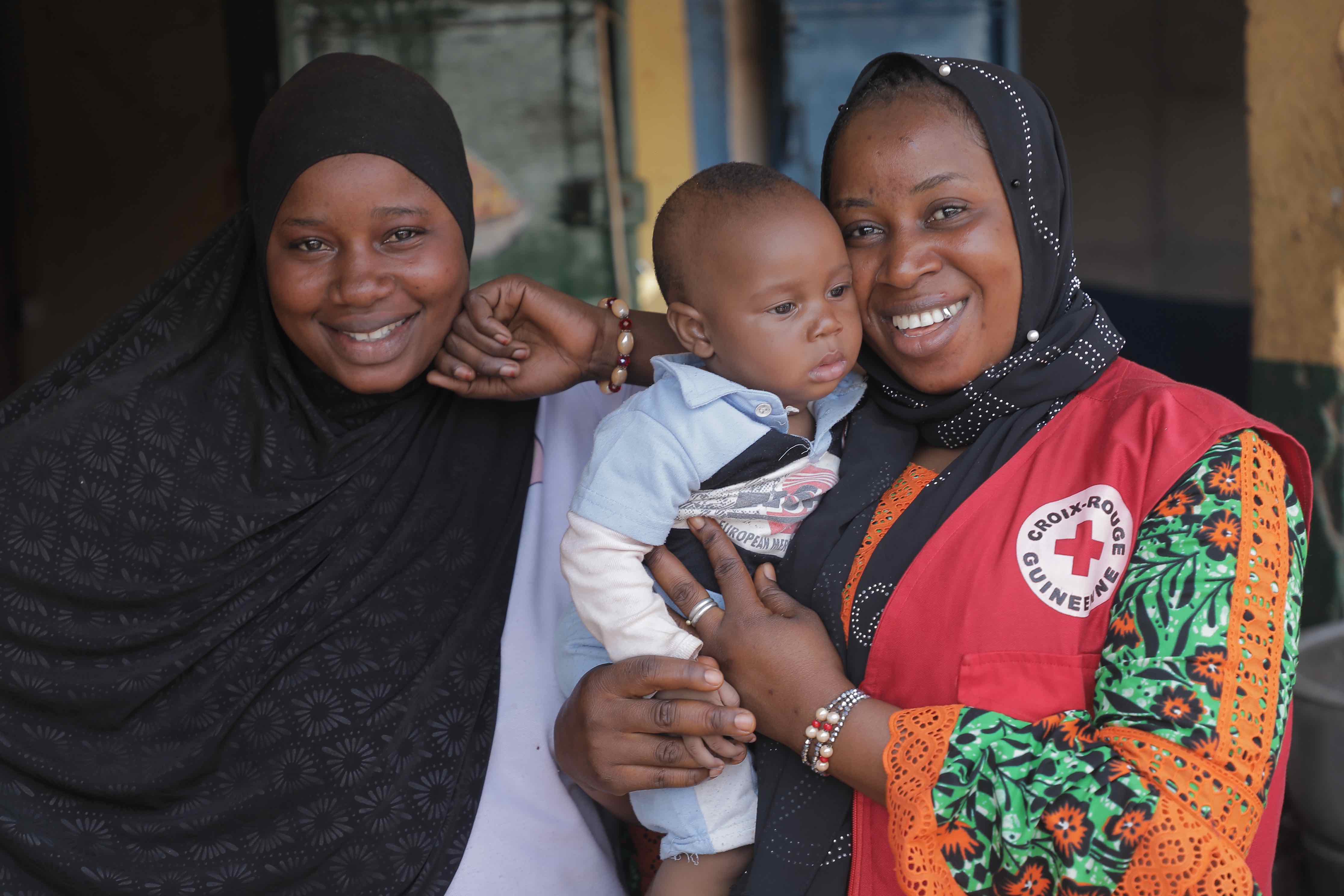 Bérété Diakité, a Guinea Red Cross supervisor with the Community Epidemic and Pandemic Preparedness Programme, holds a baby boy who she helped get vaccinated after sensitively engaging his mother (left) on the benefits of childhood immunization.