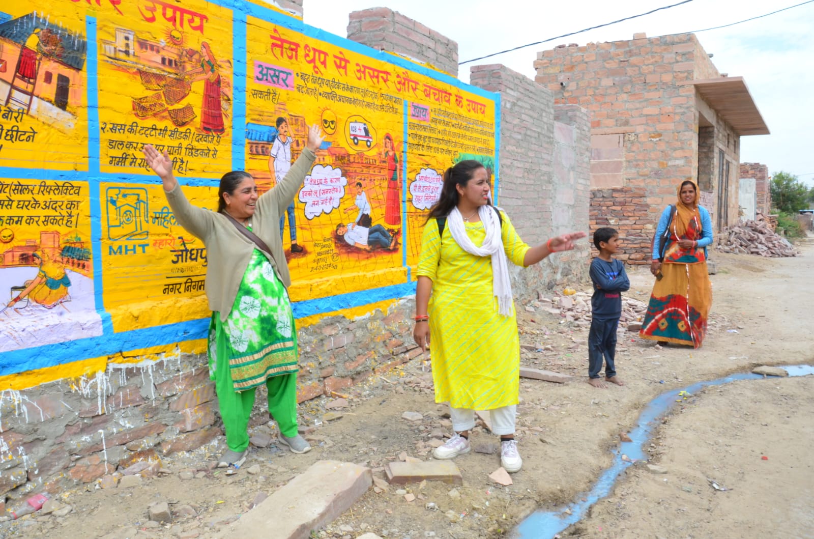 Tres mujeres y un niño frente a un mural creado por Mahila Housing Trust en Jodhpur (India). El mural forma parte de una iniciativa más amplia para mitigar los efectos del calor extremo en las comunidades vulnerables de Jodhpur mediante sistemas de alerta temprana y sensibilización.