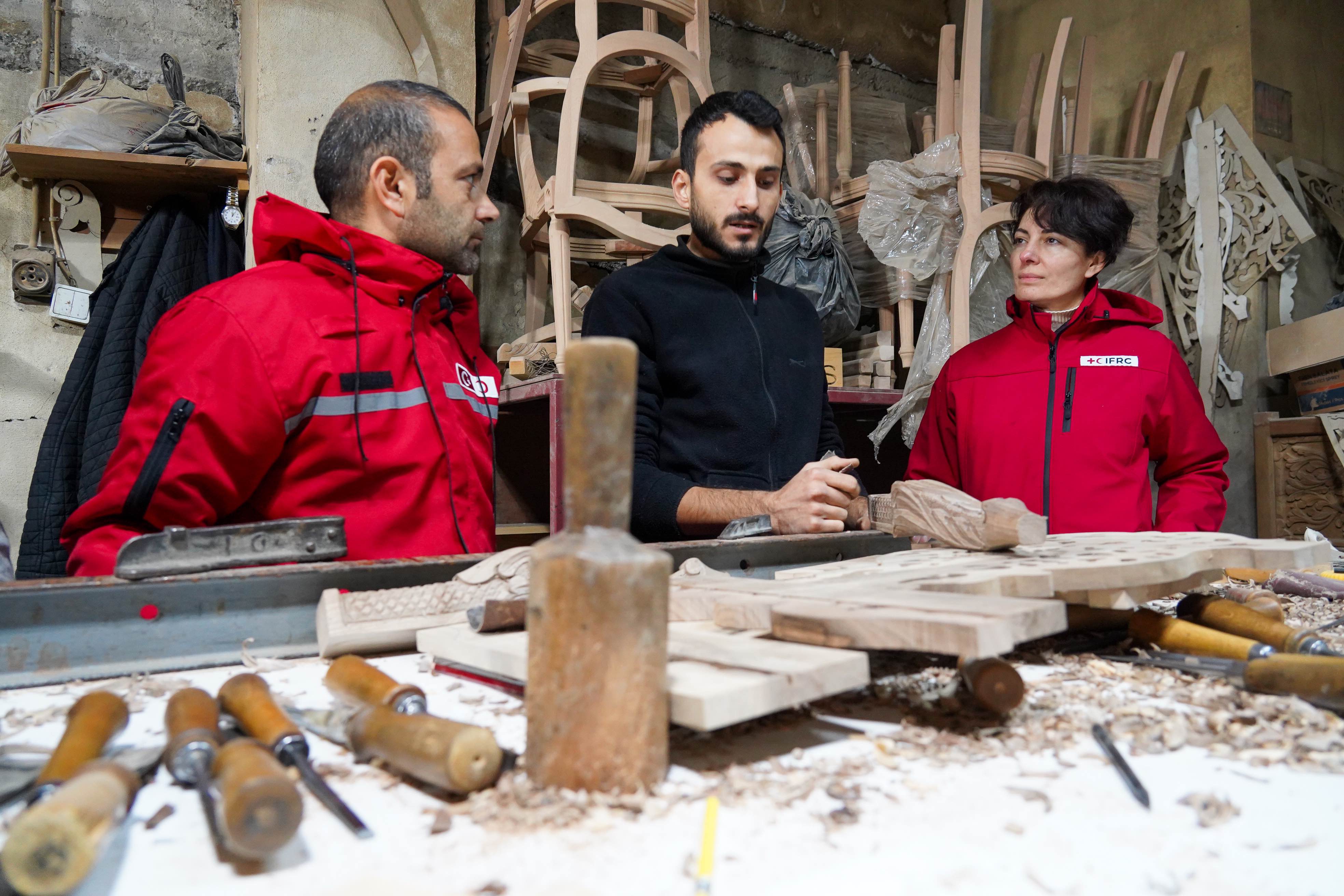 A team from the Turkish Red Crescent and the  IFRC  looks on as carpenter Ökkeş İnce stands in front of his workbench, which is covered with wood and woodworking tools. İnce’s house and workshop in Kahramanmaraş were badly damaged during the earthquake.