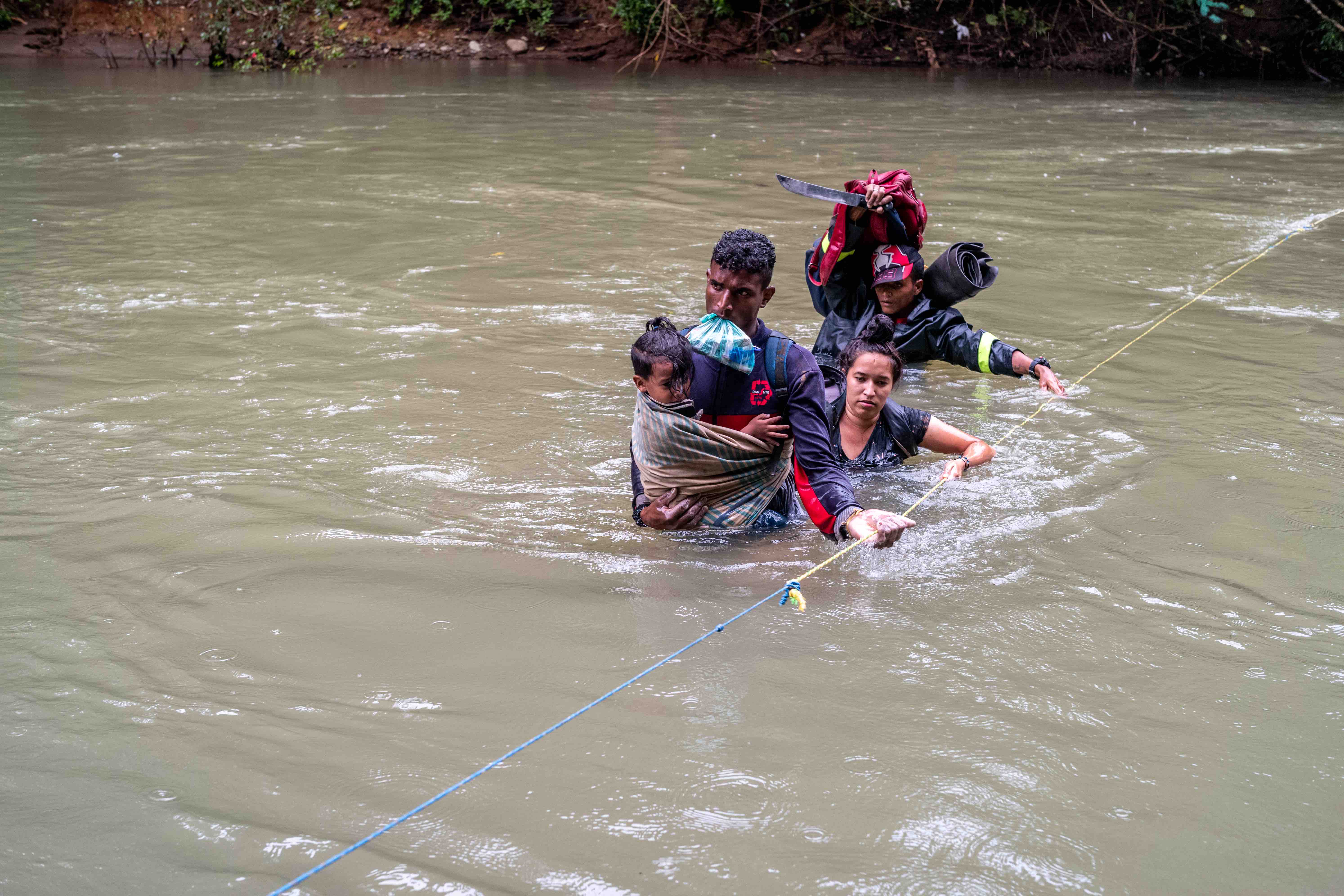 A group of migrants travers a river as they grip a cable that guides them and helps keep them on track despite the rushing current.