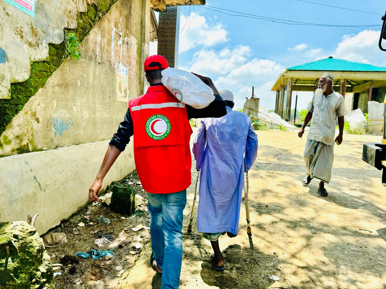 A Bangladesh Red Crescent volunteer helps a man, walking with crutches due to missing leg, to carry supplies back to his home.