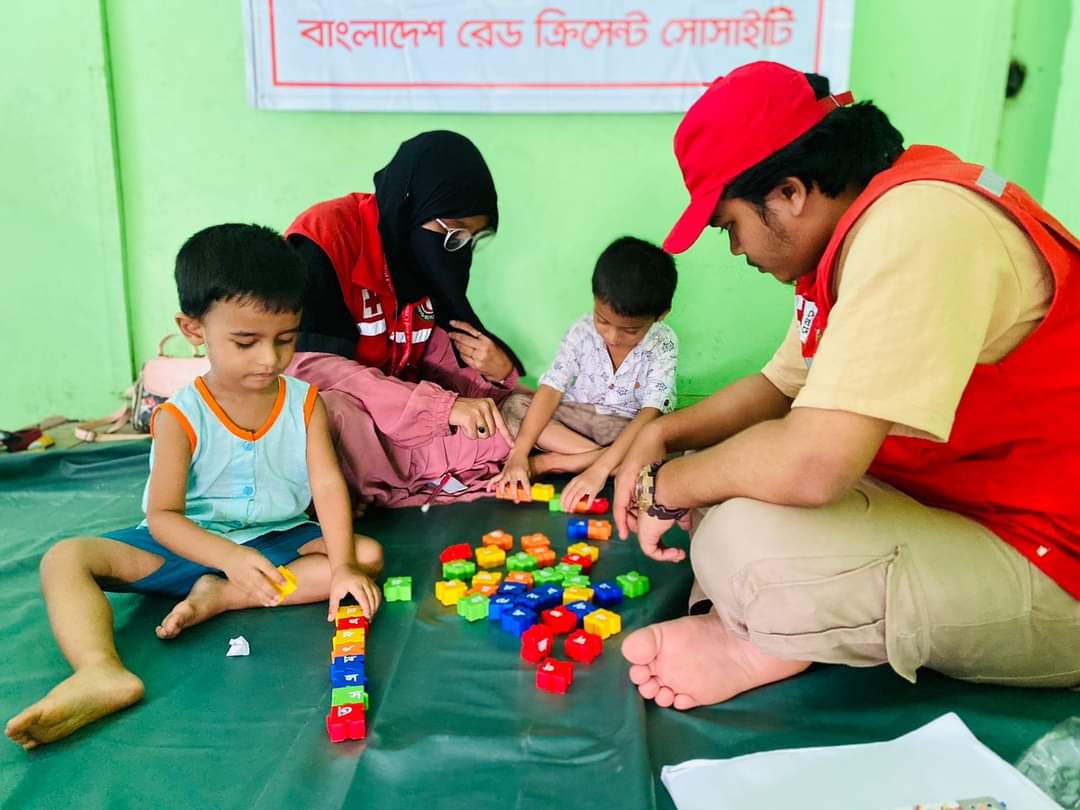 Two volunteers with the Bangladesh Red Crescent play games with two children to help take their minds off the stress of losing their homes.