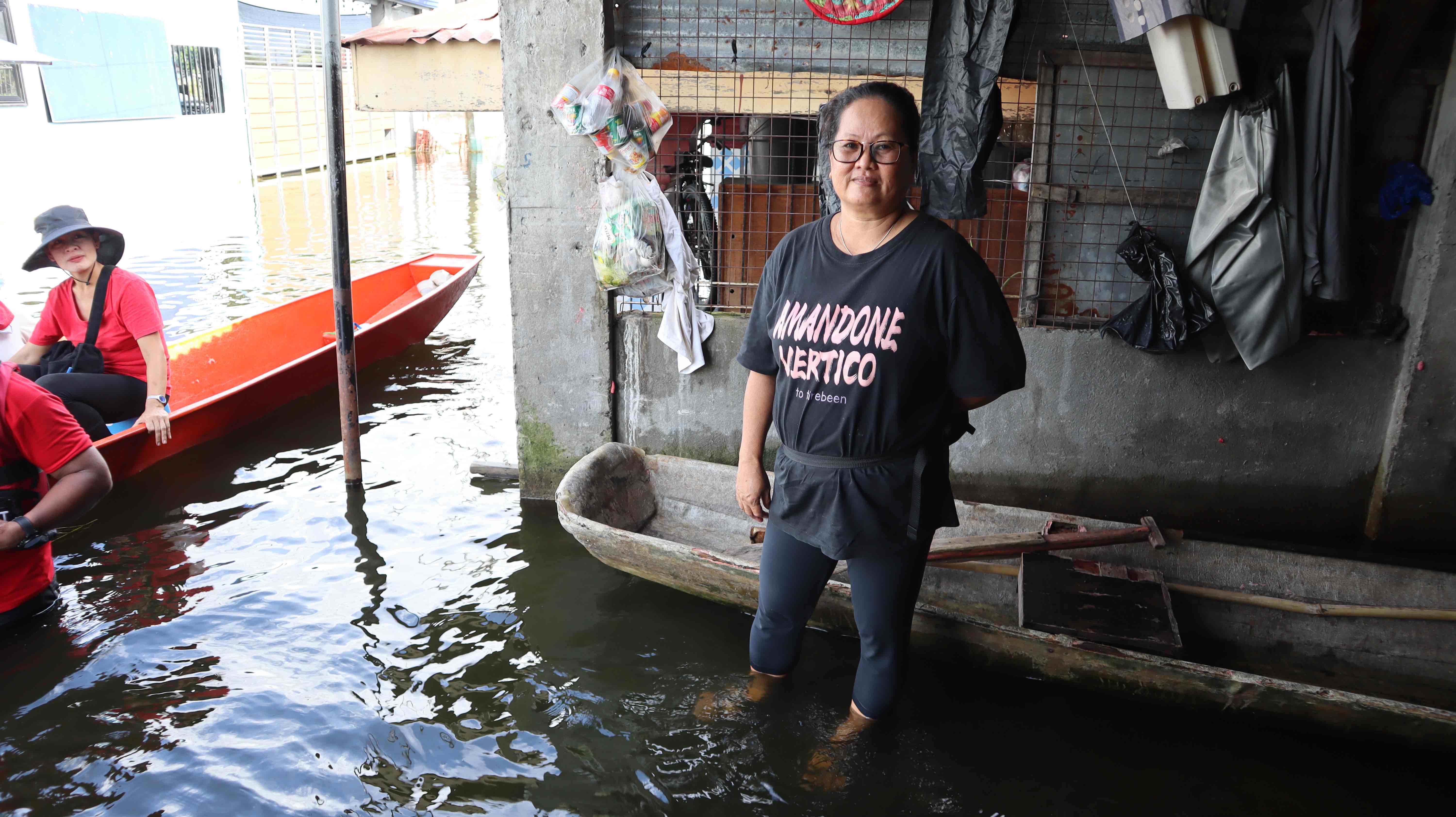 Meldrid Liodel, a 53-year-old single mother, stands outside her home as neighbors ride by in a canoe. People in Meldrid’s community have been living in ankle- to knee-deep water now for three months. This is having a significant impact on people's health.