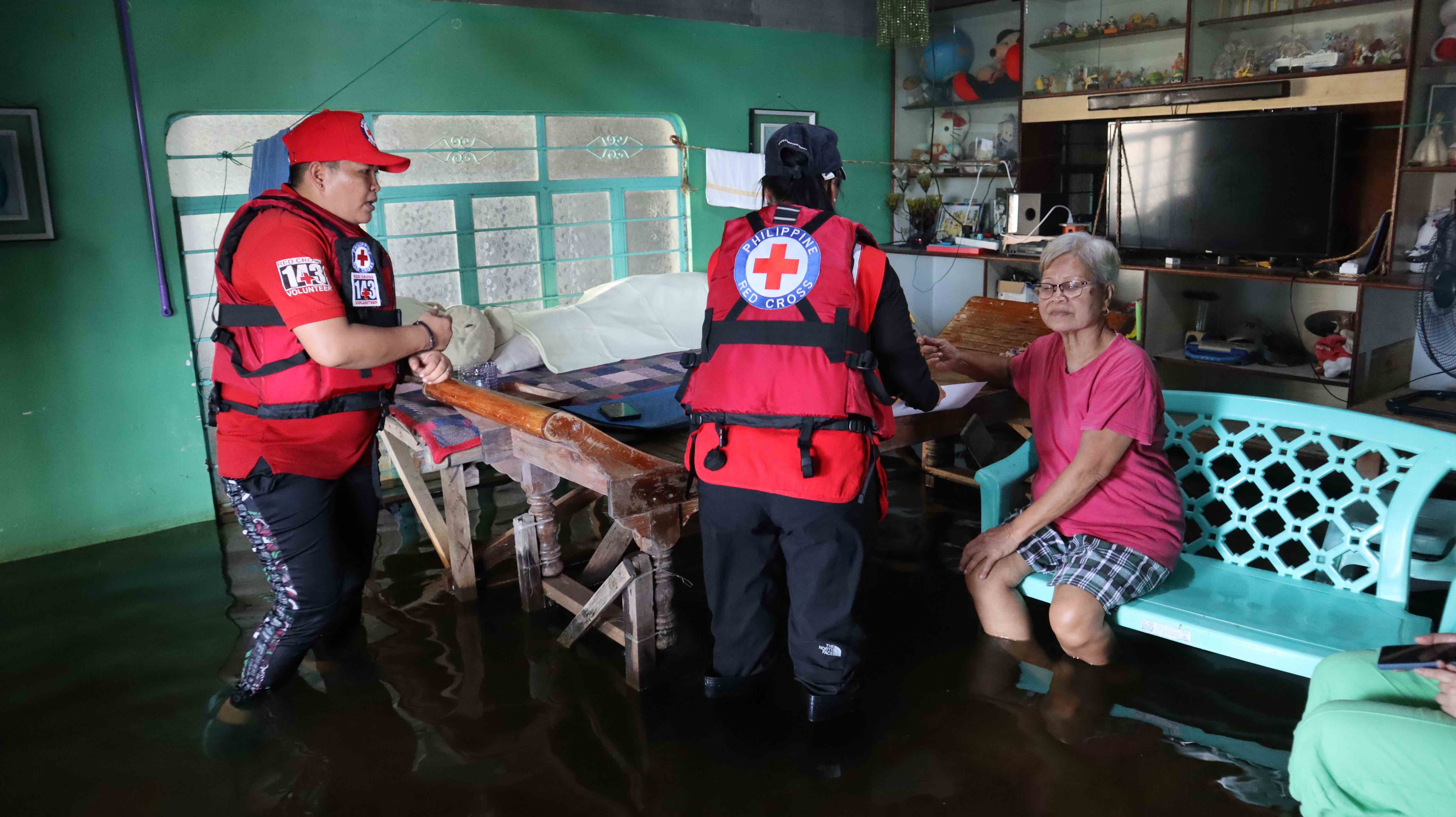 A team from the Philippines Red Cross pays a visit to Amelia Torres, who sits on a bench in her living room, her feet submerged in murky water. Her home has been carpeted in shin-deep water since July 2024.