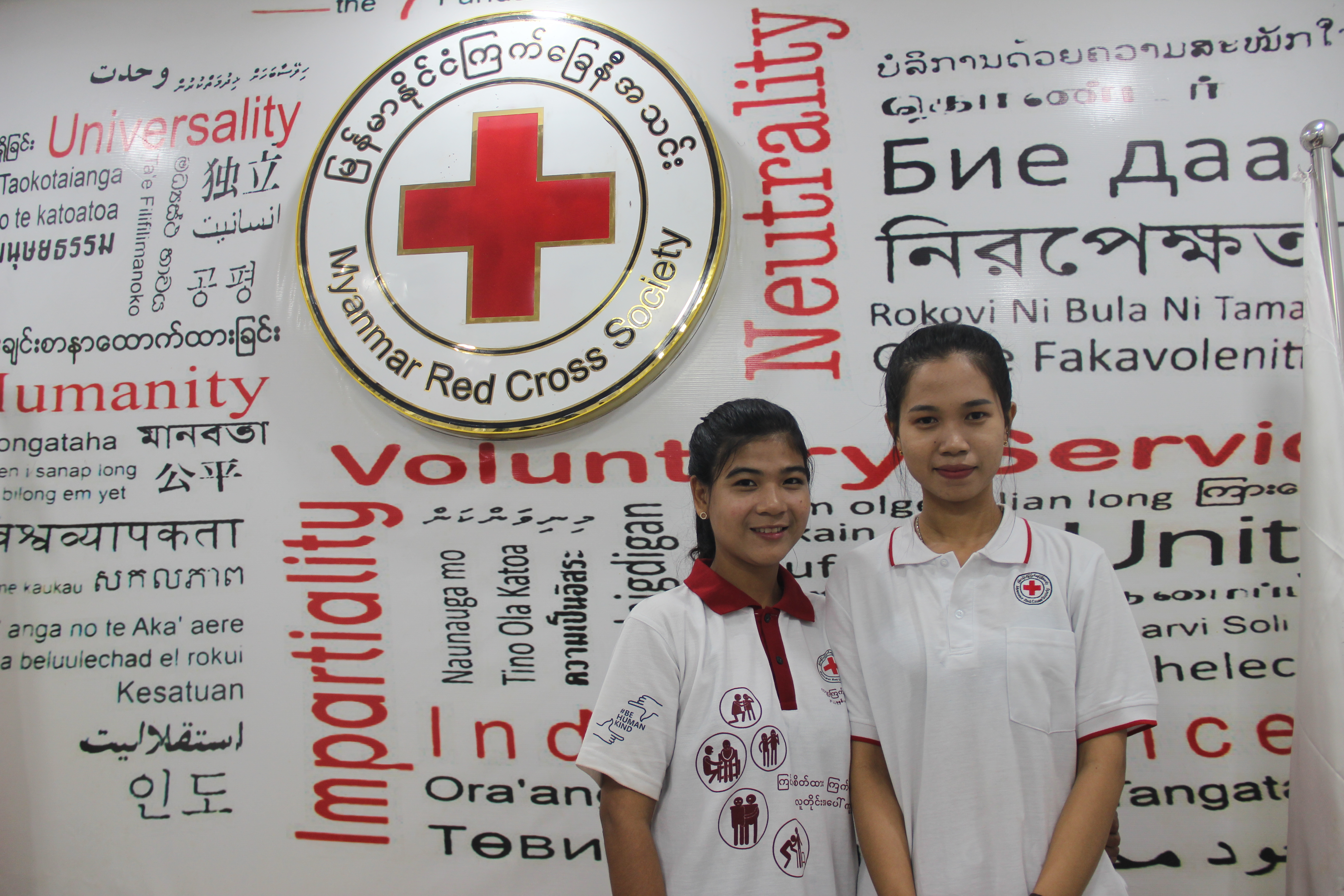 Cho Cho Aye y Phu Ngon Wai ante un muro en el que aparecen palabras que representan los principios, valores y actividades humanitarias clave de la Cruz Roja de Myanmar.