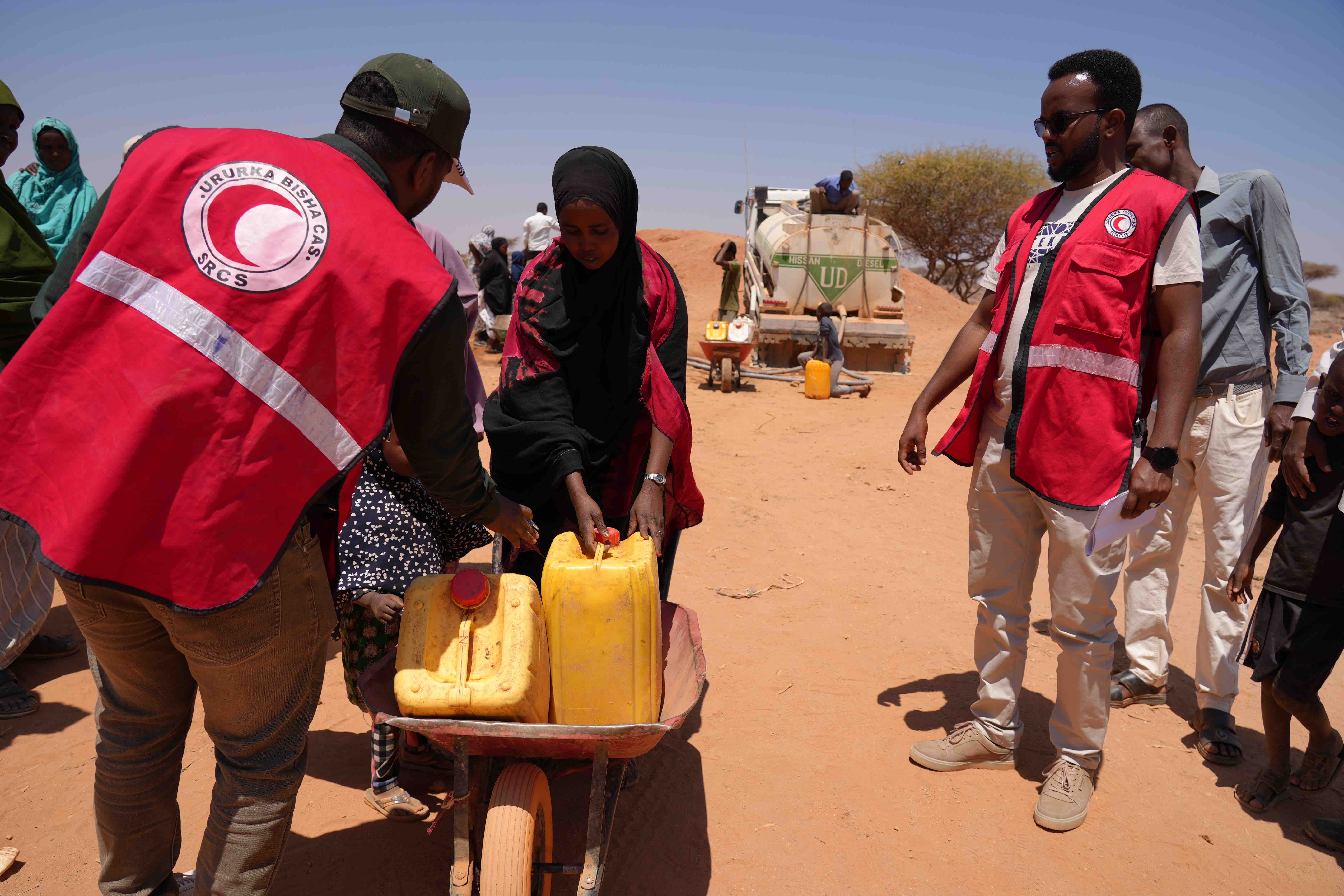 SRCS staff assist Dahir, a mother of six, in loading water onto her wheelbarrow after the weekly water truck arrives at Geed Abokor IDP camp. With no reliable water source, families rush to collect just 2–20-litre jerricans, barely enough to last until next week's delivery. 
