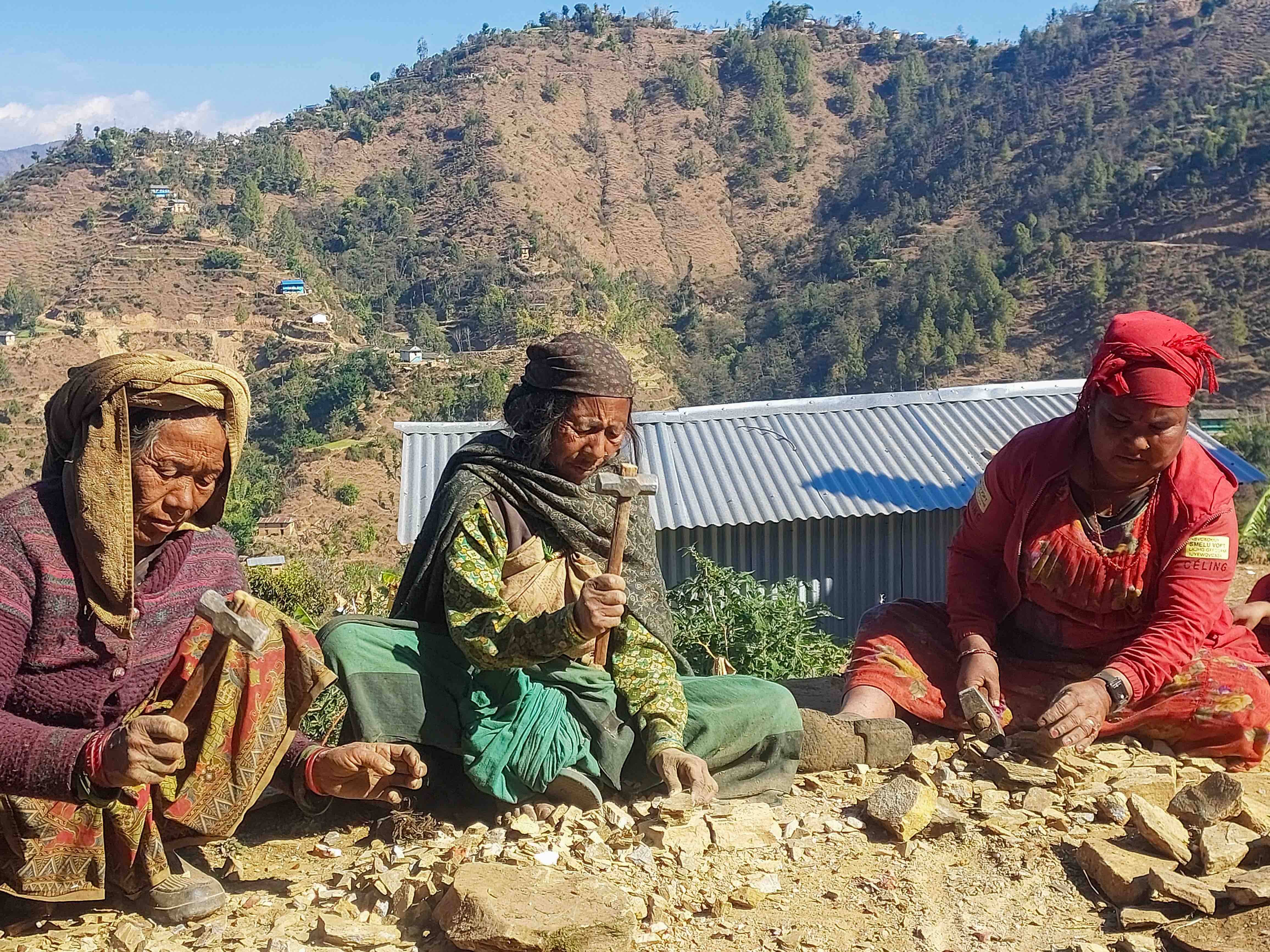 Women from Darma-6, Salyan, sit on the ground as they break small rocks into gravel that will be used in the construction of household water taps.
