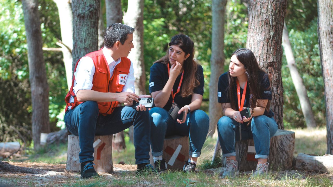 Andrés Morales conversa con dos jóvenes durante el campamento de la Cruz Roja Uruguaya en marzo de 2025. Las tres personas se sientan en troncos de madera al aire libre. 