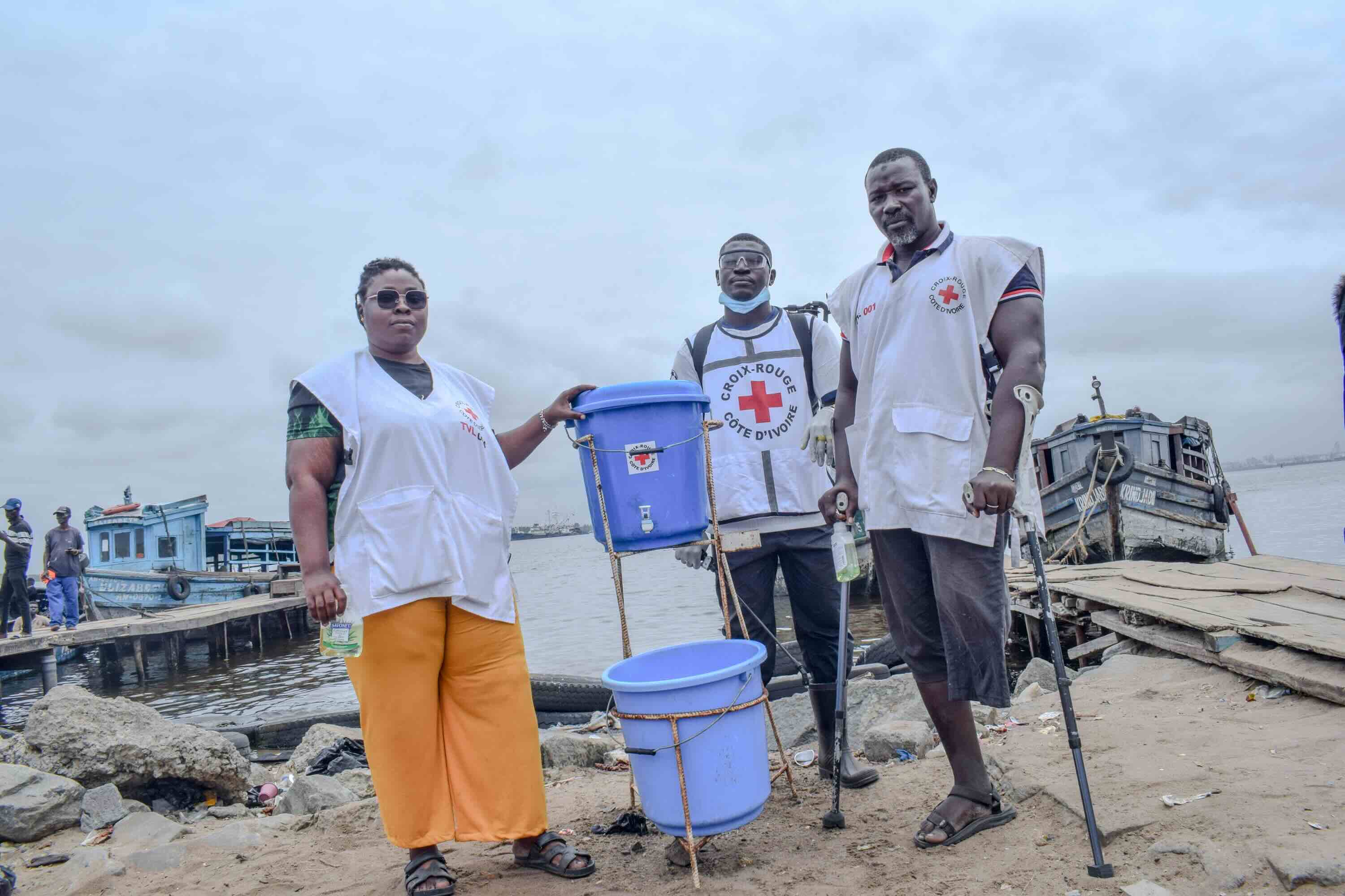 Des volontaires de la Croix-Rouge de Côte d’Ivoire se tiennent près d’un dispositif de lavage des mains qu’ils ont installé sur le rivage, là où les gens prennent le ferry pour traverser un lagon non loin de la capitale, Abidjan. 