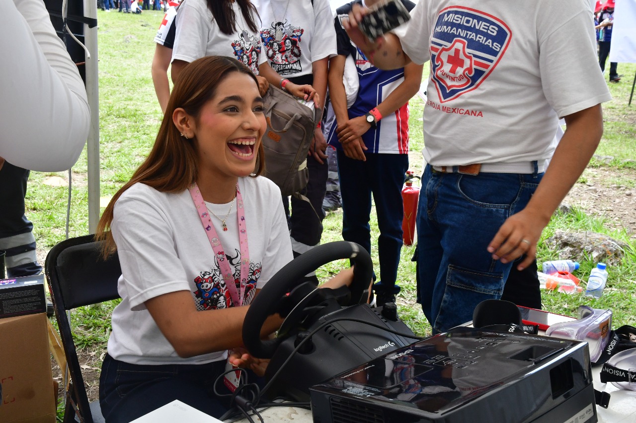 Una joven voluntaria sonríe mientras participa en una actividad con un simulador de conducción, rodeada de otras personas jóvenes en el campamento nacional de juventud de la Cruz Roja Mexicana, en agosto de 2025.