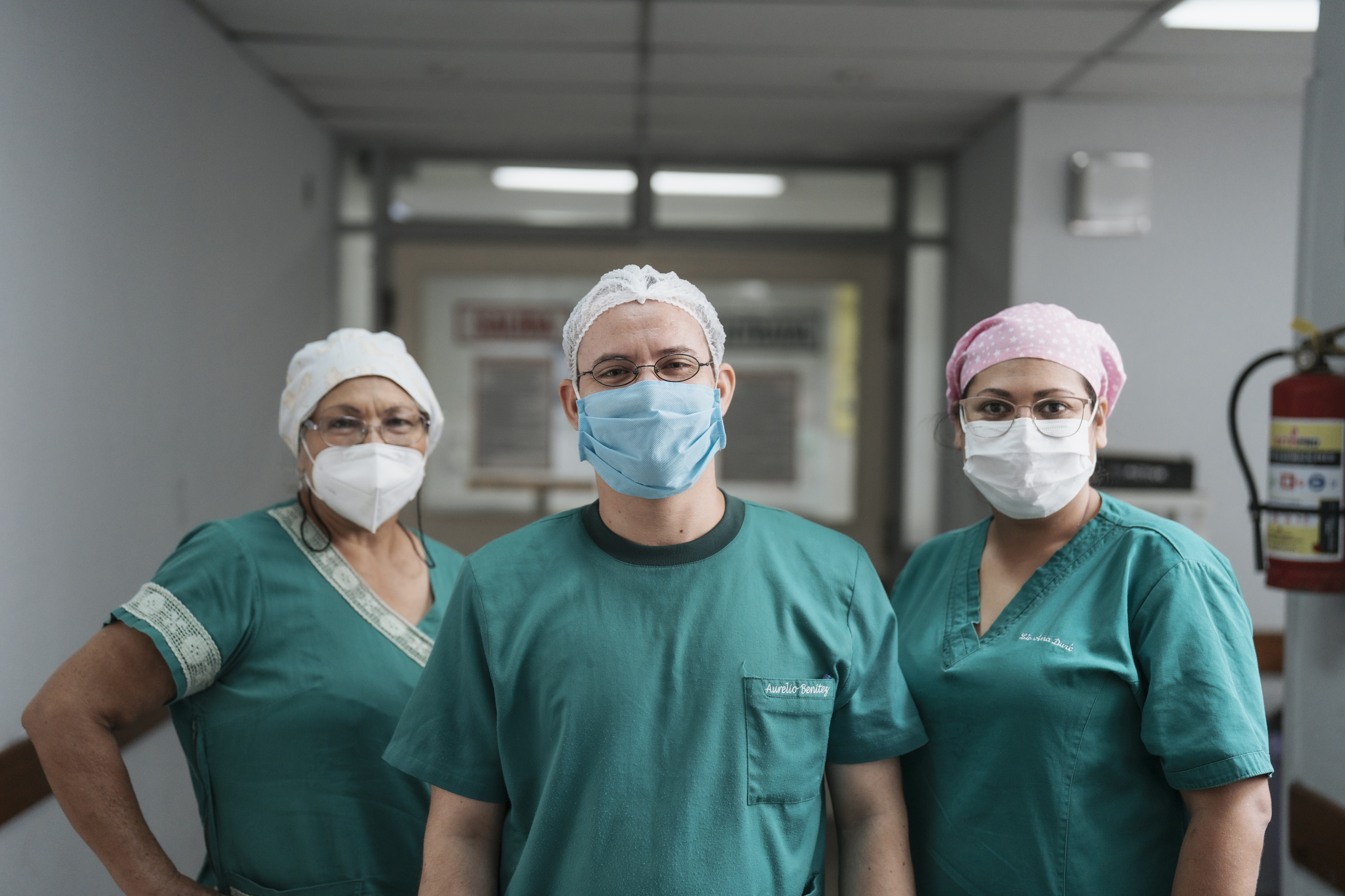 Three members of the Reina Sofía Hospital healthcare staff pose in a hallway, smiling through their medical masks and uniforms.