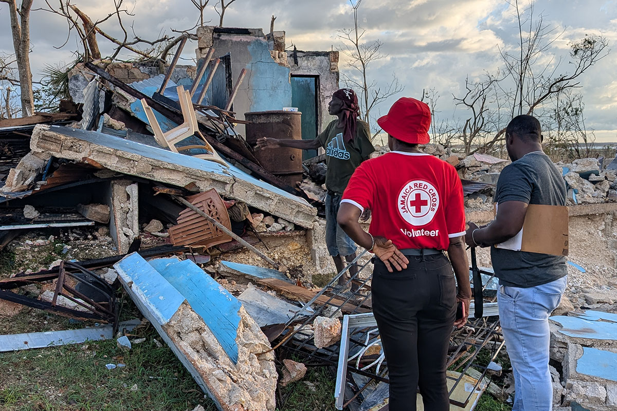 A Jamaica Red Cross team and a local resident stand beside a home destroyed by Hurricane Melissa in November 2025, as teams deployed across St. Elizabeth to carry out damage and needs assessments and distribute emergency supplies.