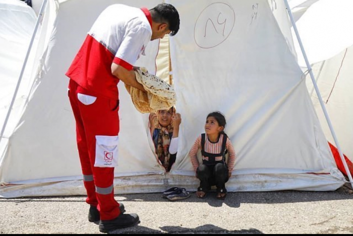 Two young girls sits by a tent for internally displaced people in Iran and are offered bread by an Iranian Red Crescent volunteer