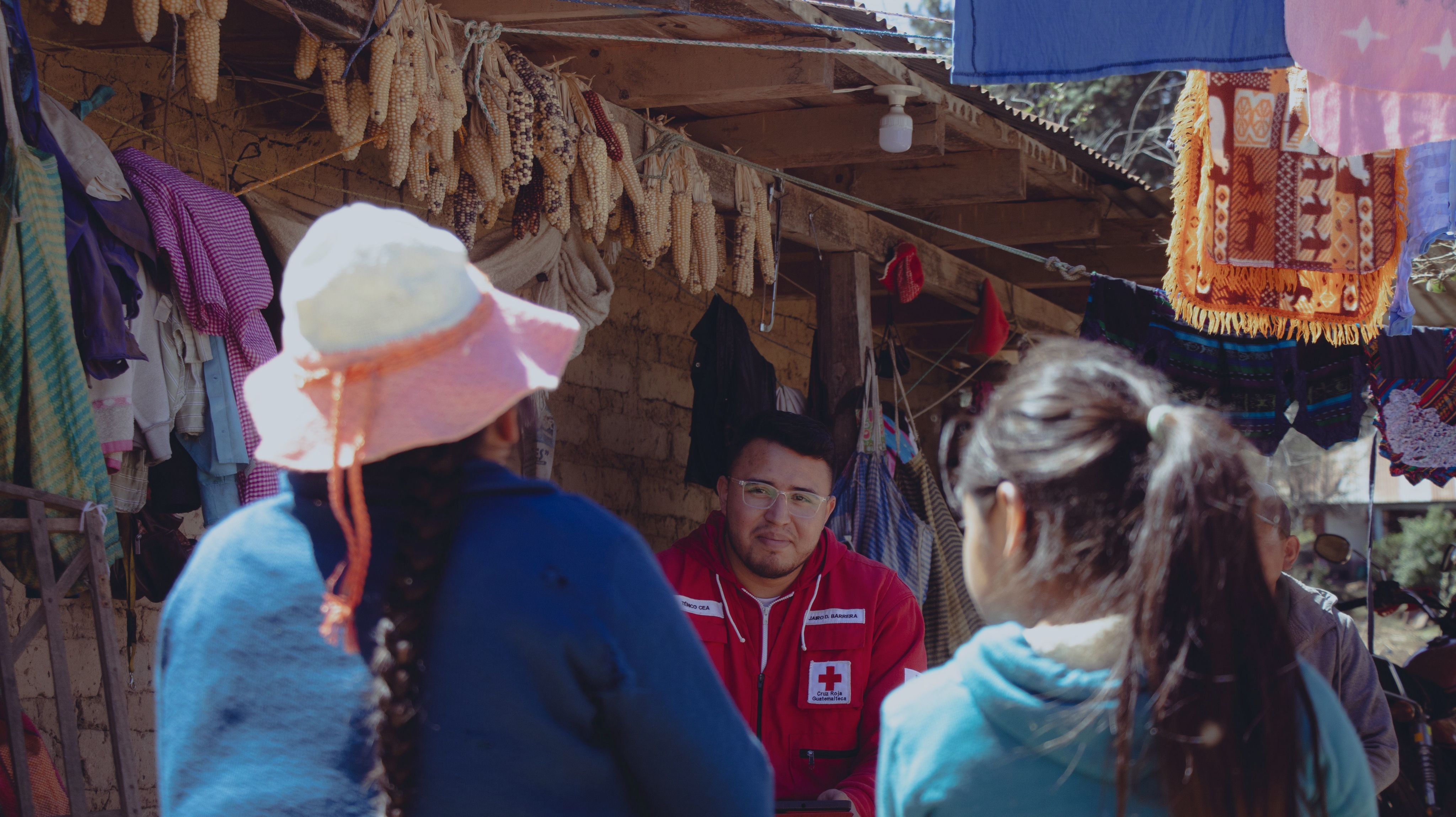 Es un plano medio, en el centro hay un voluntario de Cruz Roja Guatemalteca que ve a la cámara, al frente hay dos mujeres, una en cada lado de la imagen. La de la izquierda usa sombrero y una larga trenza.La de la derecha es una niña con una cola larga 