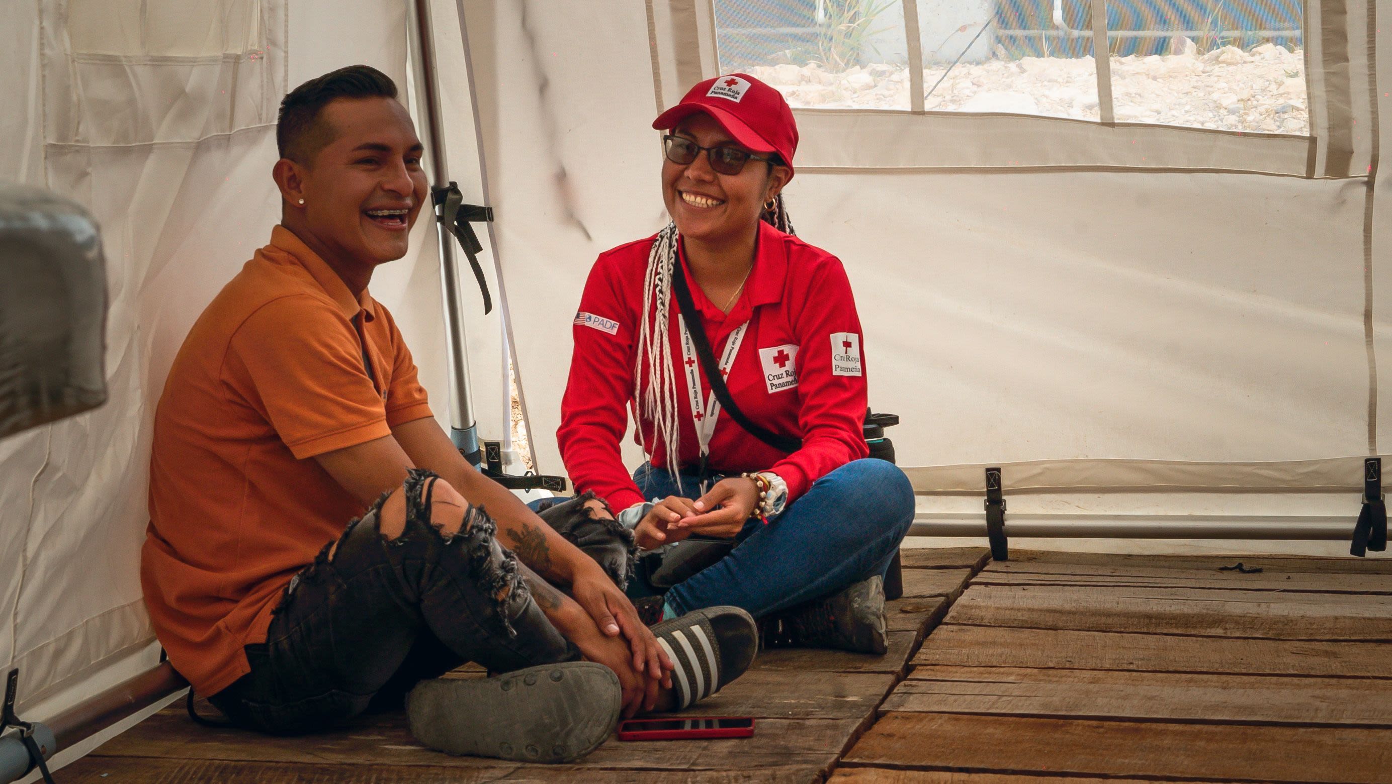 Una voluntaria de la Cruz Roja Panameña está sentada en el suelo junto a un joven migrante dentro de una carpa en un campamento en Darién. Ambos sonríen