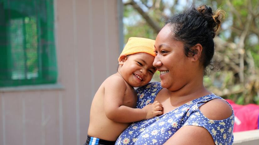 A mother in Tongatapu, Tonga stands with her baby boy nestled against her shoulder as she receives shelter assistance from Tonga Red Cross volunteers.