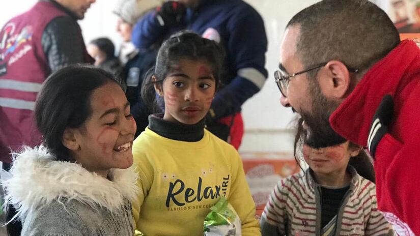Voluntario de la Media Luna Roja Palestina distribuyendo alimentos mientras comparte risas con una niña afectada por el terremoto en Siria.