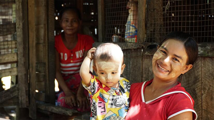 Mother Moe Thuzar stands holding her baby, 'Lucky Boy', outside her home in Mon State, Myanmar, with her own mother in the background.