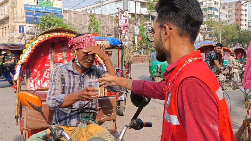 متطوع في الهلال الأحمر البنغلاديشي يقدم كوبًا من الماء لسائق عربة في نارايانغانج، بنغلاديش في يونيو/حزيران 2023 لمساعدته على التأقلم مع الحرّ الشديدة.