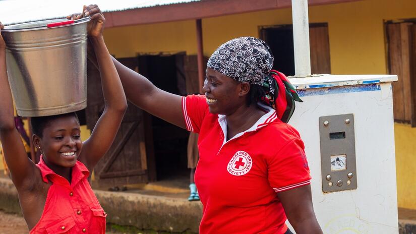 Regina Nyamevor, a volunteer from the Ghana Red Cross, helps a girl balance a bucket of water she's collected from the pump on her head to take to her home.