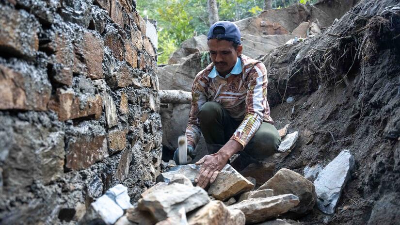 In one village, this man uses a hammer to shape rocks that will be used to build the wall of a water basin at a new water distribution point in rural eastern Nepal.