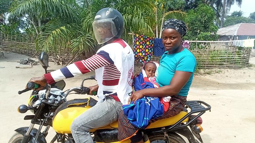 Le volontaire Joseph, Baindu et le bébé Senesie sont assis ensemble sur la moto de Joseph.