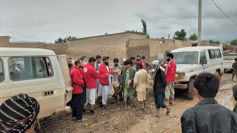 A group of people, including some from the Afghan Red Crescent Society, gather near two vehicles in a muddy street, affected by floods