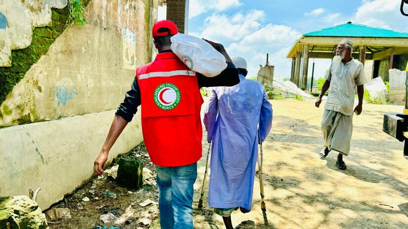 A Bangladesh Red Crescent volunteer helps a man, walking with crutches due to missing leg, to carry supplies back to his home.