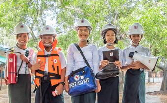 A group of five students in Kim Chaung village, Myanmar pose for a photo while wearing emergency equipment - helmets, fire extinguisher, life jacket, flashlight, and a first aid kit bag - provided by the American Red Cross to help them be prepared for disasters.