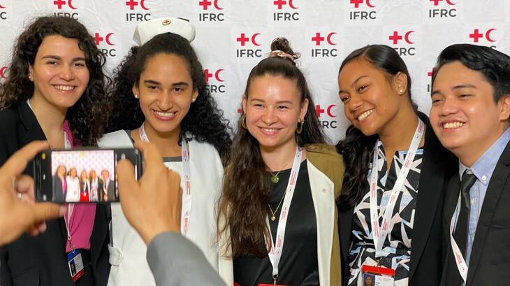 Jóvenes voluntarios y voluntarias de la Asamblea General 2022 sonríen y posan para una foto delante de un muro fotográfico con la marca de la IFRC.