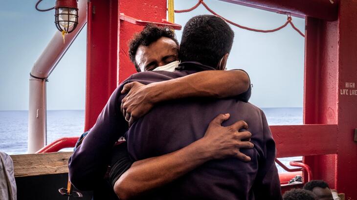 A man rescued in the Central Mediterranean by SOS MEDITERRANEE rescue teams in late June 2022 tightly hugs a fellow survivor on board the Ocean Viking rescue ship. IFRC teams also on board provide humanitarian assistance to those rescued as they wait to disembark safely on land.